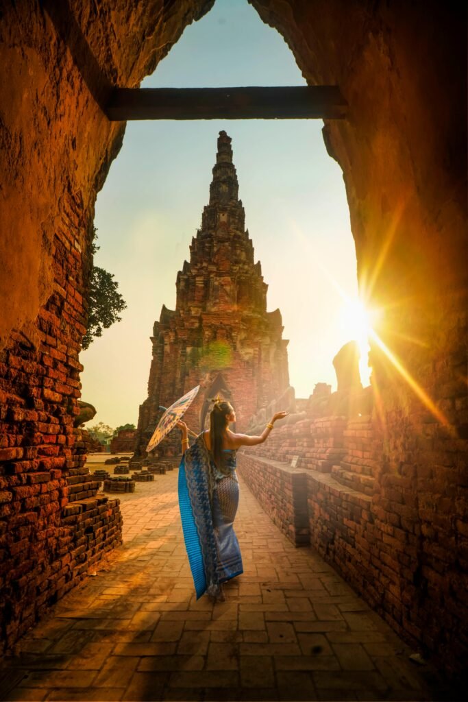 A woman in traditional wear stands with an umbrella at a historic Ayutthaya temple during sunset.